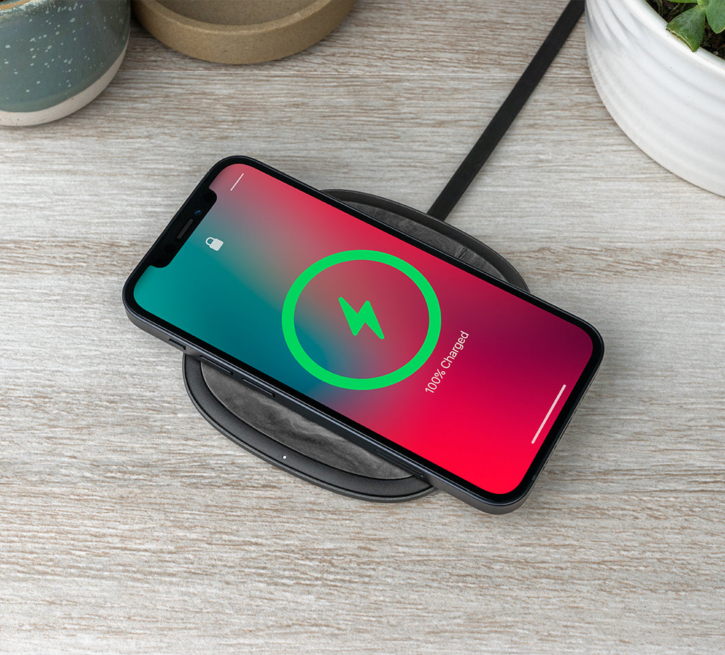 Smartphone fully charged on a wireless charging pad on a light wooden surface with plants nearby.