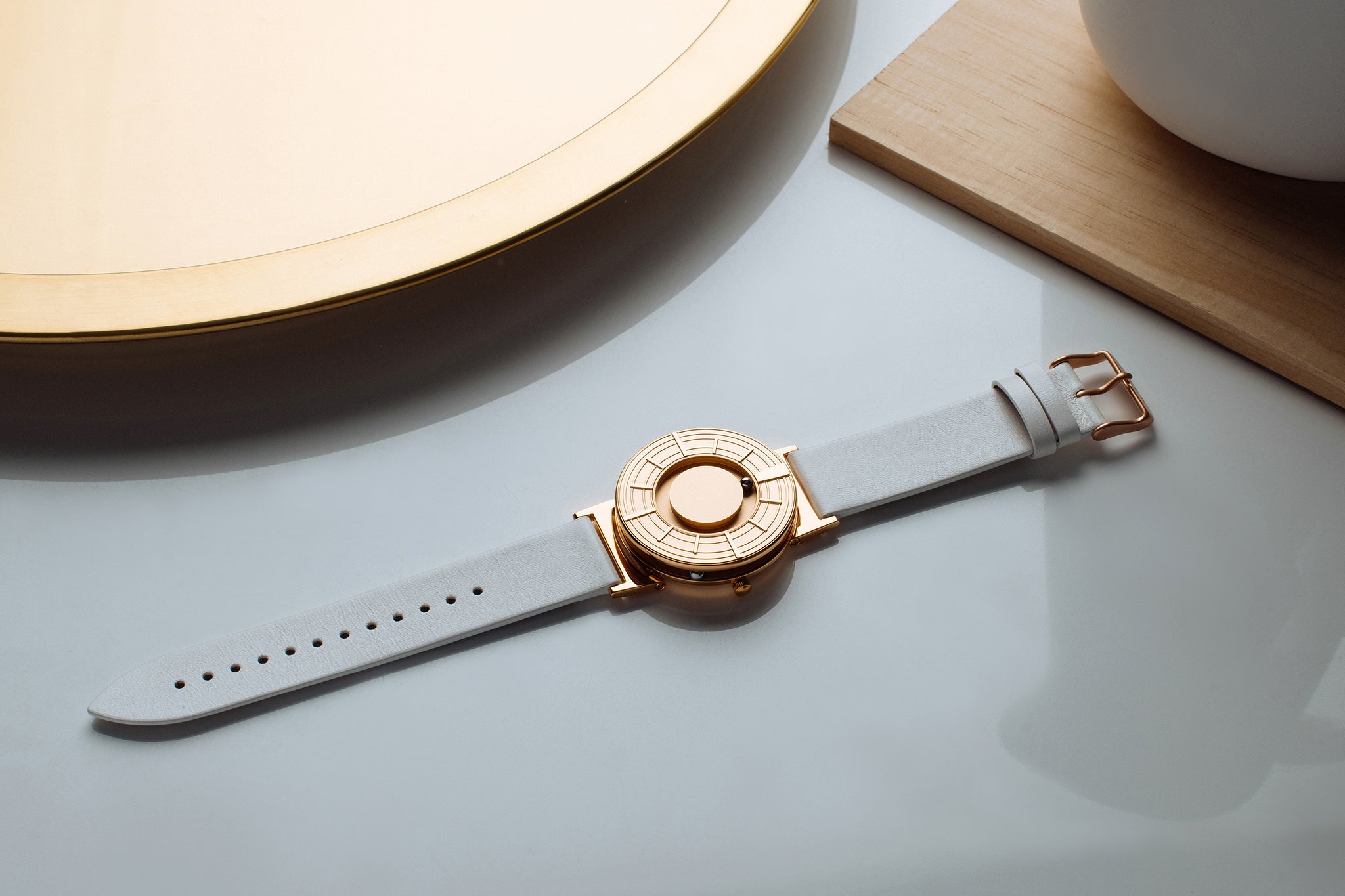 The Bradley Edge Rose Gold is flat on a white glossy surface. There is a round gold disk in the top left corner. There is a square gold object in the top right corner.