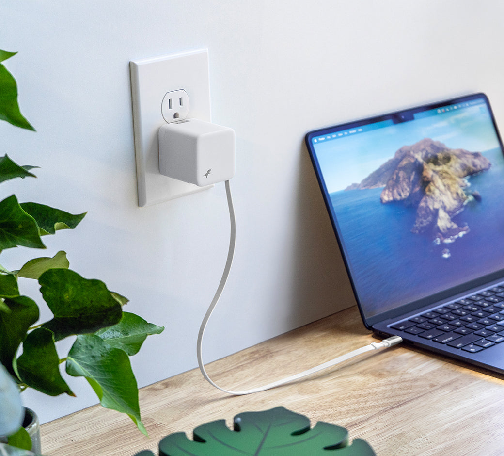 White wall charger with USB-C cable connected to Macbook on desk near window and small plants to the left.