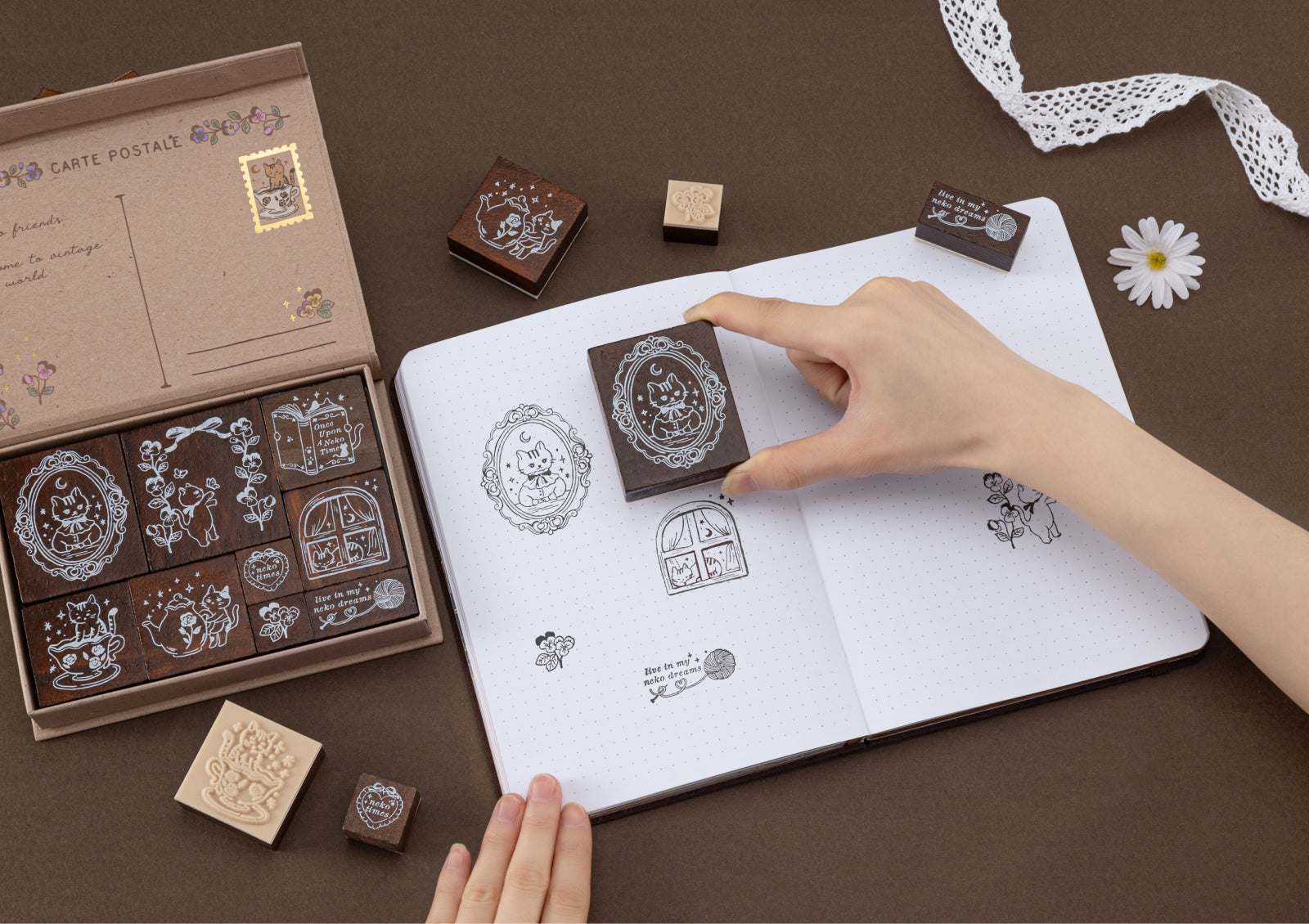 A close-up shot of a hand pressing a wooden cat stamp onto a dotted journal page, surrounded by other stamp impressions and the open product box.