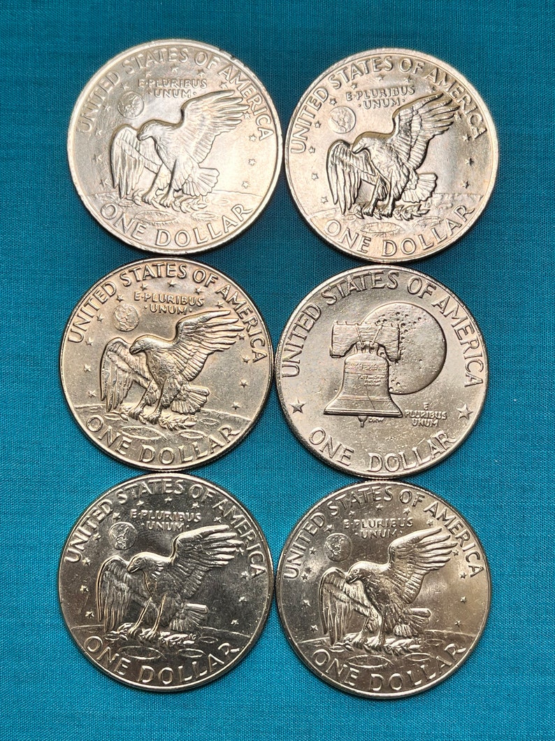May include: Six silver United States one dollar coins. The coins feature the words 'United States of America' and 'E Pluribus Unum' around the edge. The coins feature different designs in the center. Three coins feature an eagle with its wings spread. Two coins feature a bald eagle with its wings spread. One coin features a bell with the words 'Liberty' and '1776' on it.