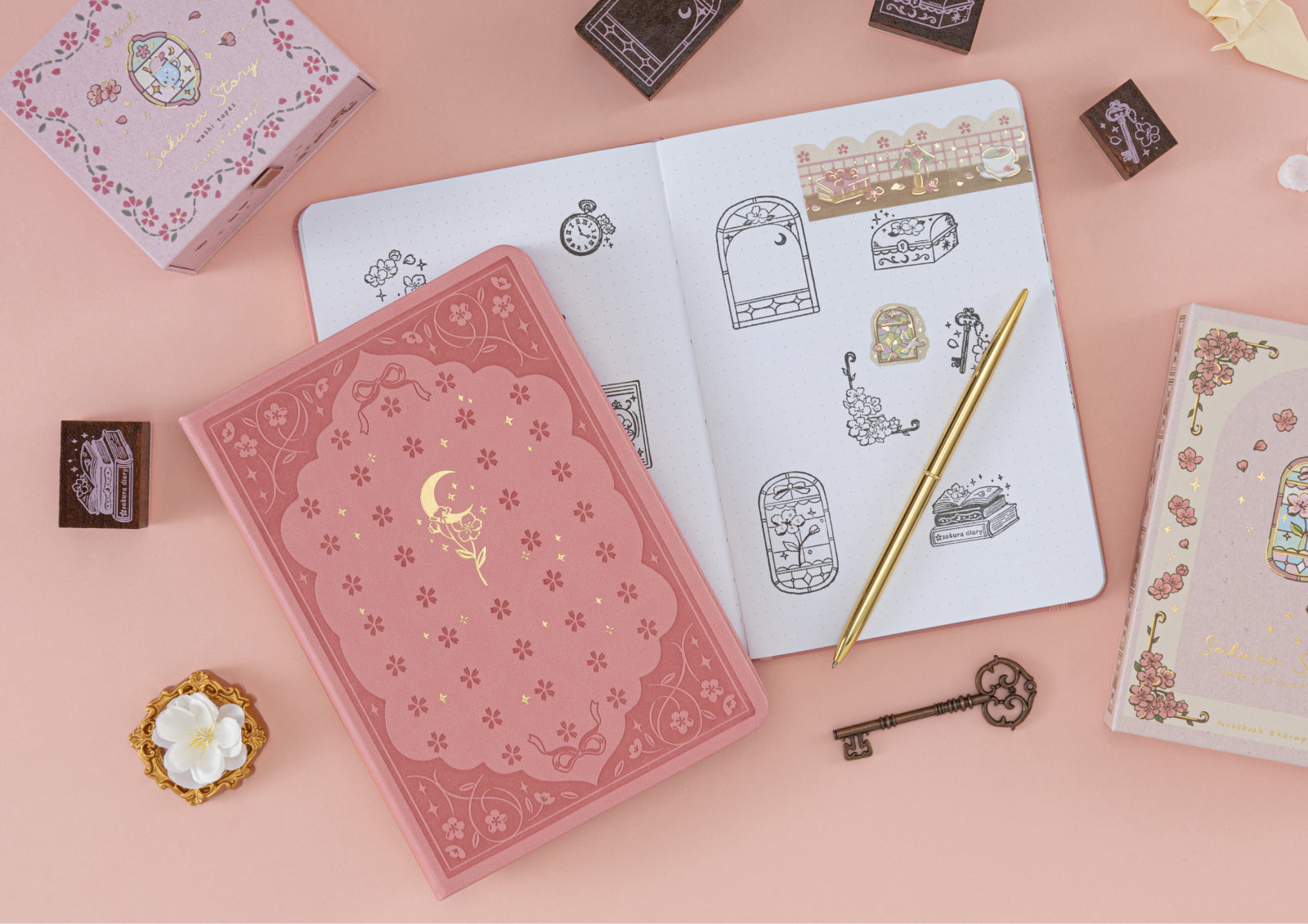 The Sakura Story stamp box and a pink floral journal displayed together with a gold pen and stationery accessories.