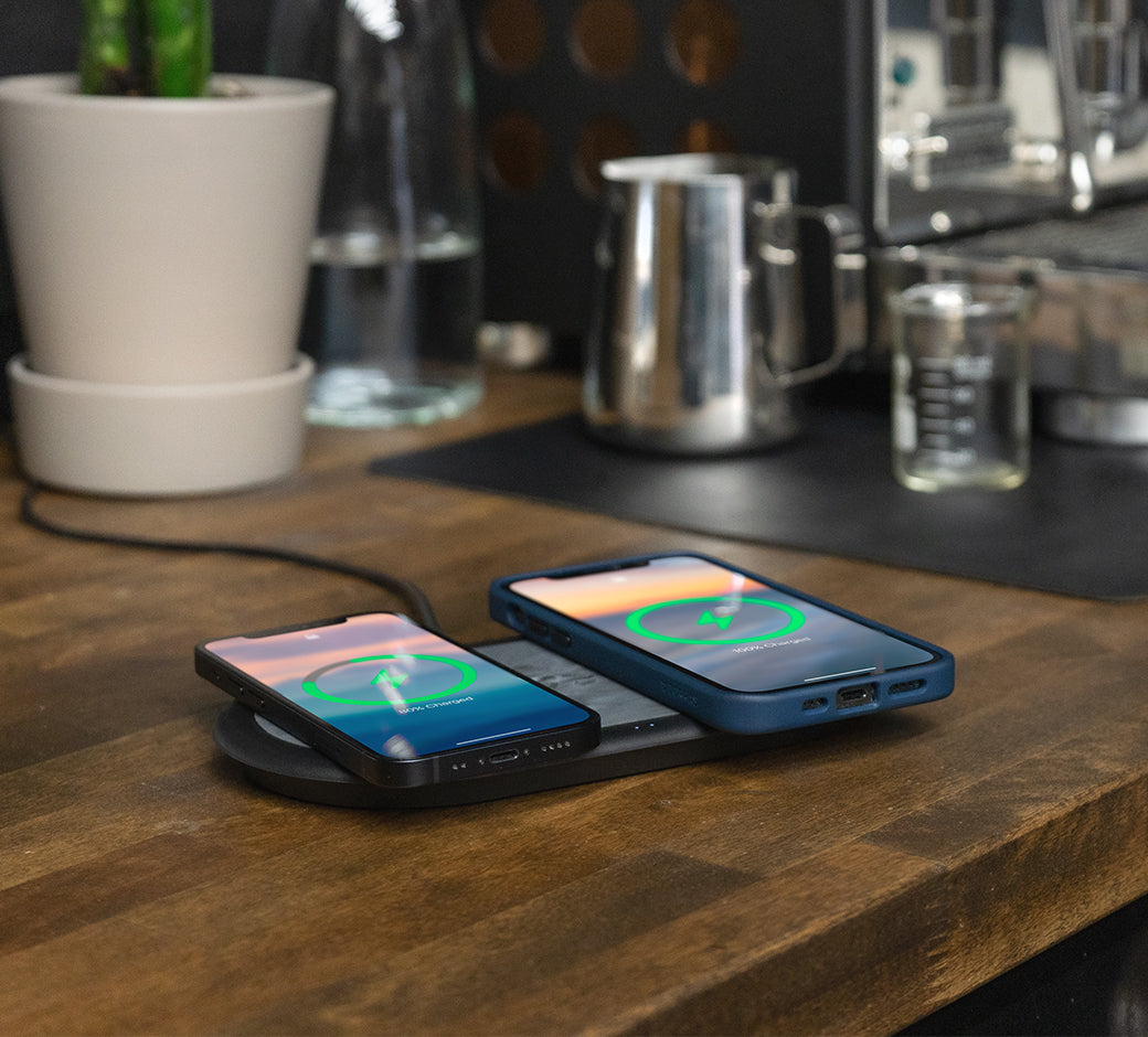 Two phones wirelessly charging on a black pad on a wooden countertop with kitchen items in the background.