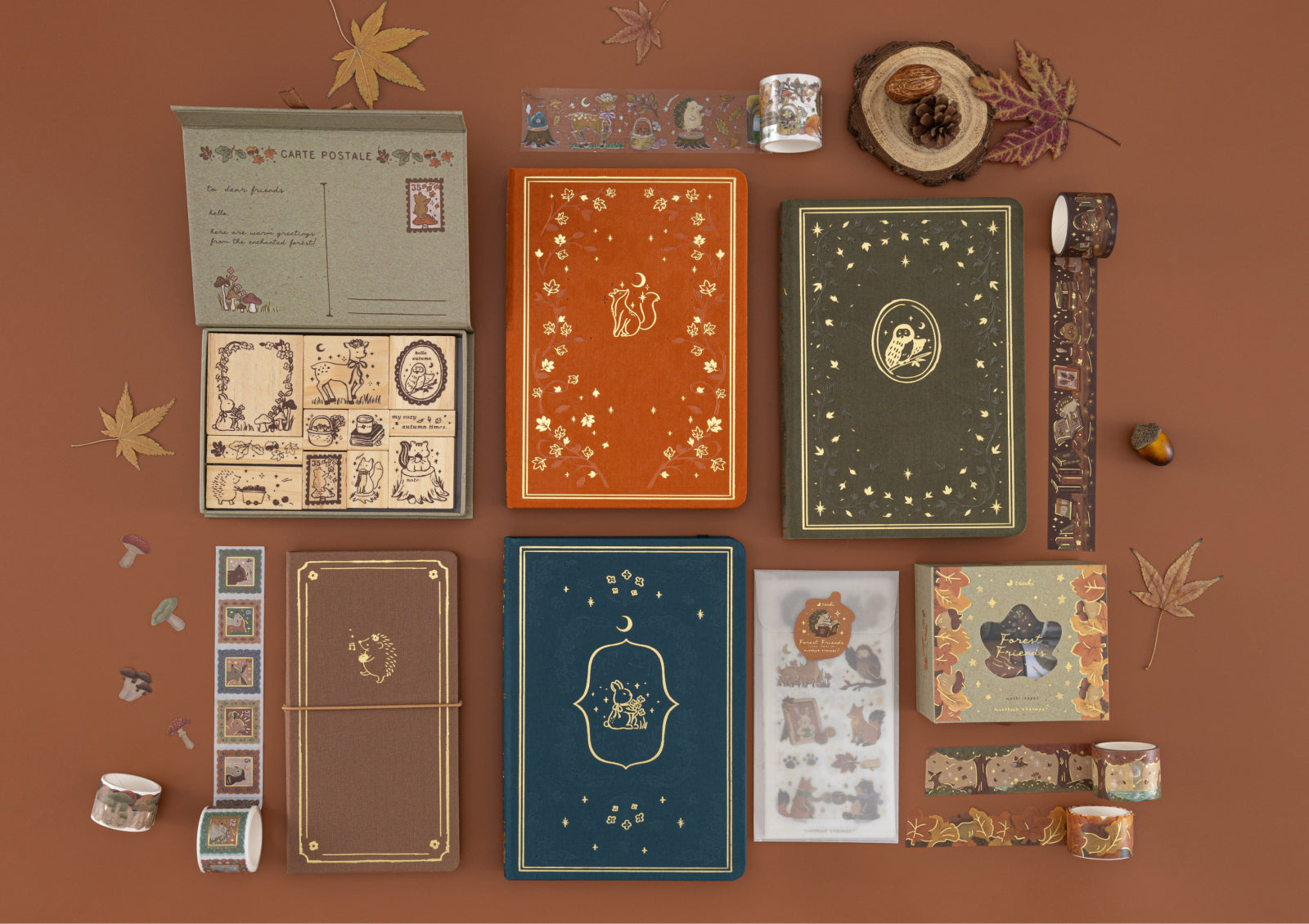 Flatlay of the full Tsuki Forest Friends collection, including four notebooks, wooden stamp box, washi tapes, and sticker set arranged with autumn leaves.
