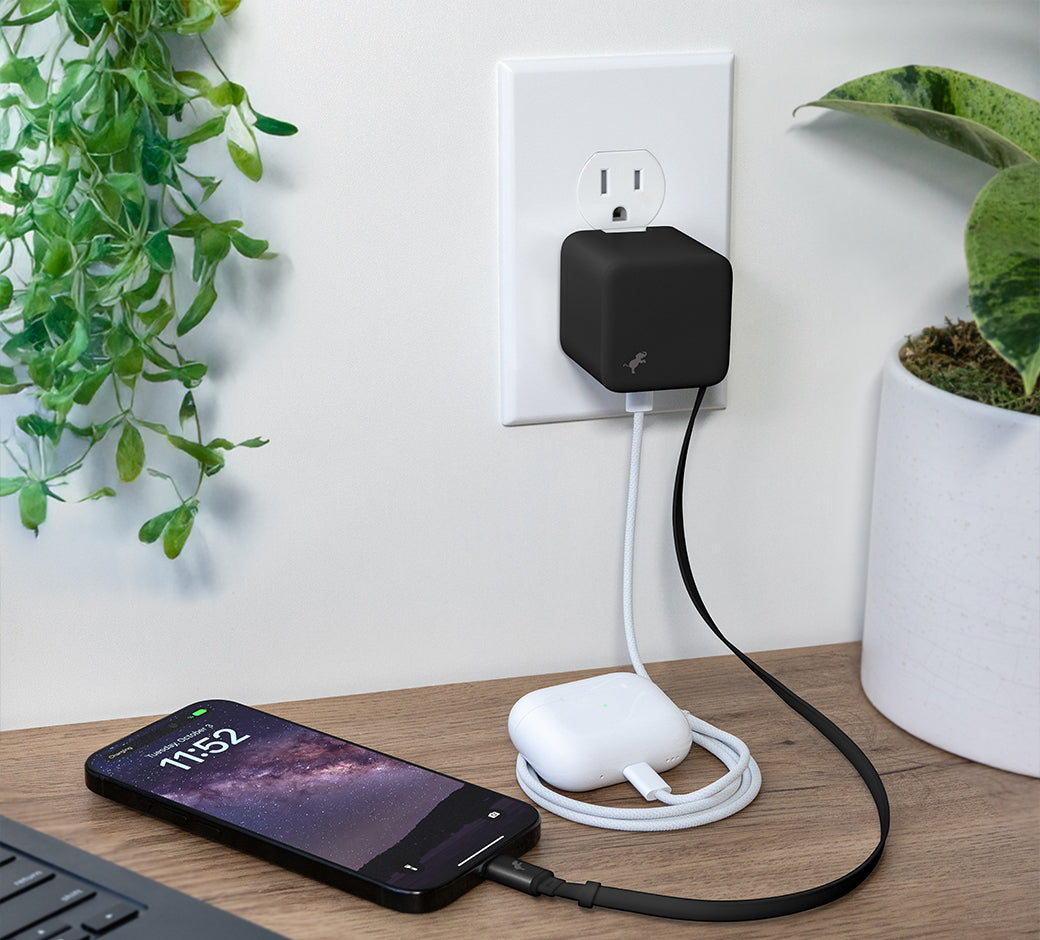 Black Wall Charger charging iPhone and AirPods on wood desk with plants to the left and right.
