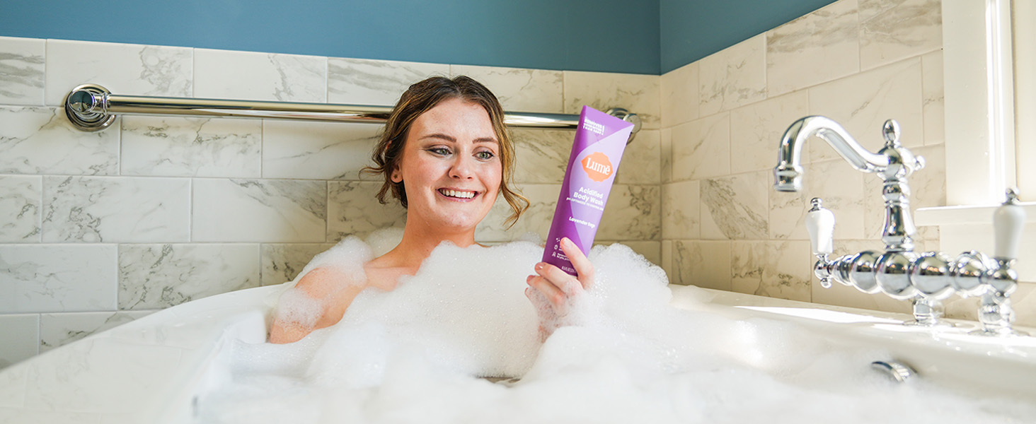 A woman using Lum� Acidified Body Wash in Lavender Sage being used in a bath tub