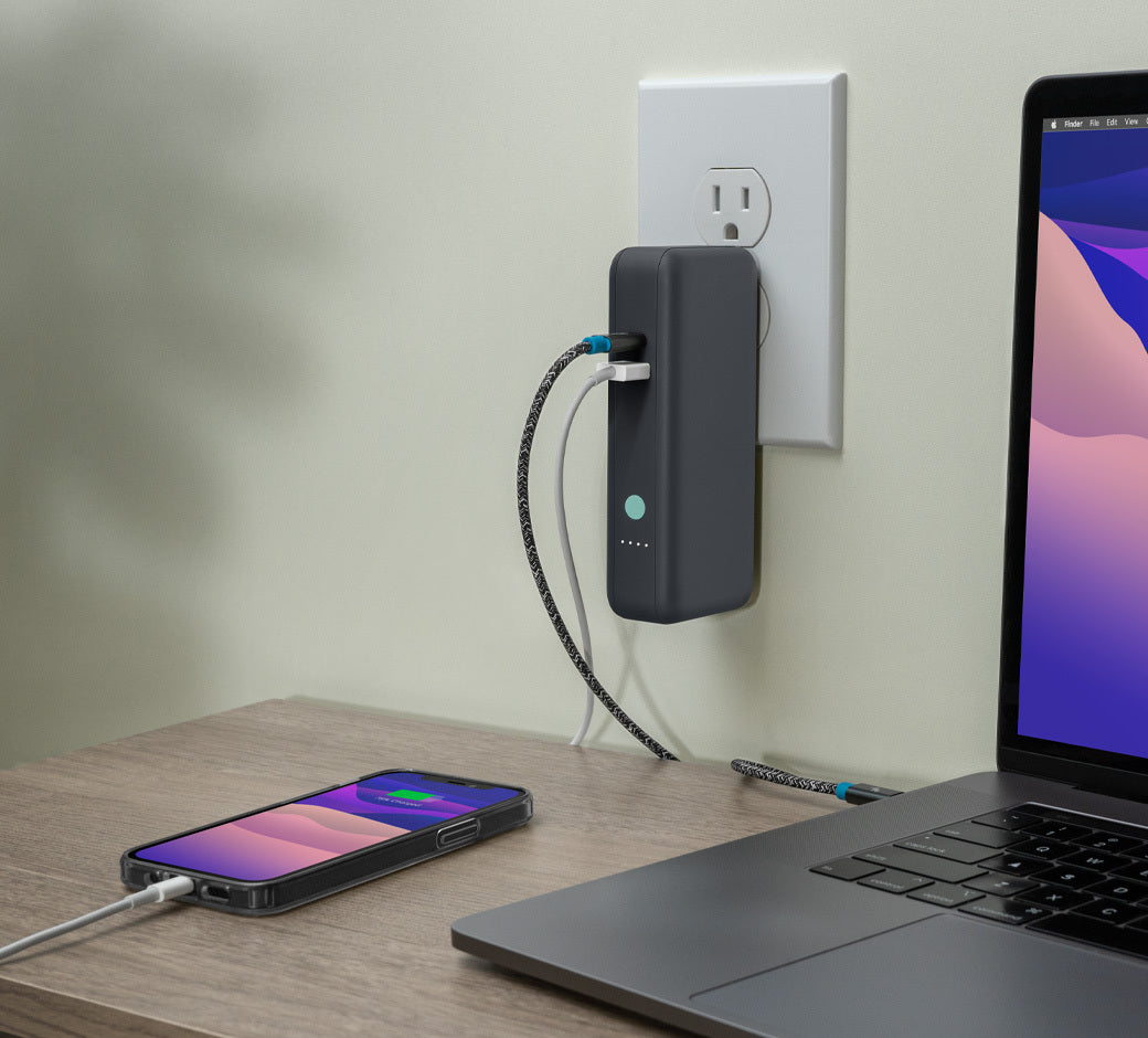 Portable power bank plugged into a wall outlet, charging a laptop and smartphone on a wooden table.