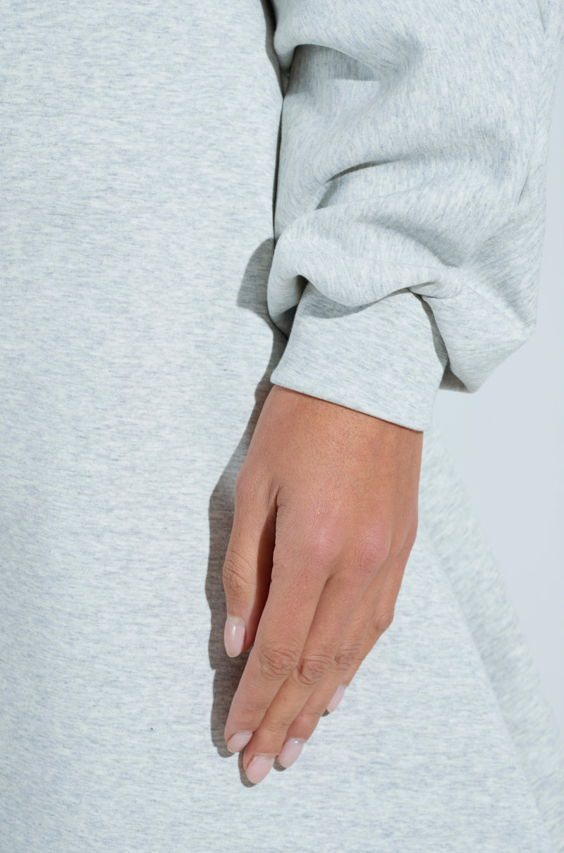 A person rests their hand against their side, dressed in a light gray COMFY COZY SWEATSHIRT JUMPSUIT. The warm skin tone and neatly trimmed nails highlight the simple, soft texture of the fabric, creating a focus on its comfort and elegance.