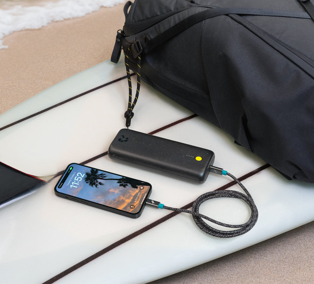 Black power bank charging a phone on a surfboard by a black backpack.