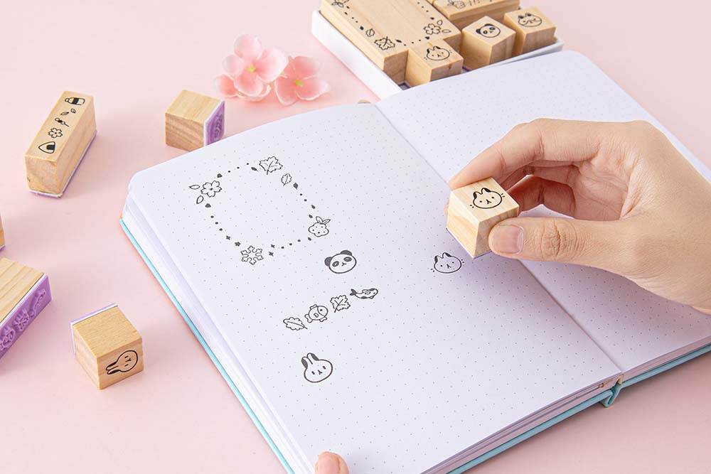 Tsuki ‘Four Seasons’ Bullet Journal Stamp Set by Notebook Therapy x Milkkoyo held in hands over open bullet journal page spread with pink flowers on petal pink background