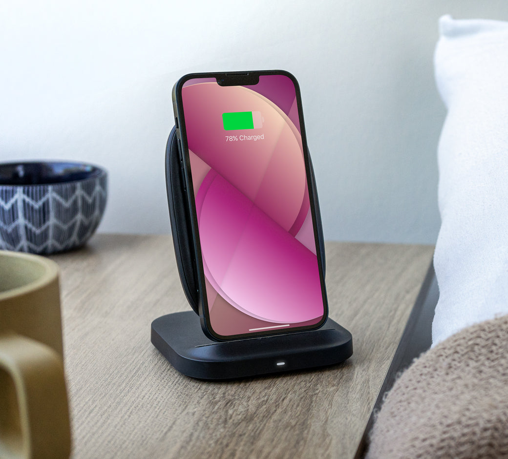 Smartphone charging on a black stand on a bedside table with cups and pillows nearby.