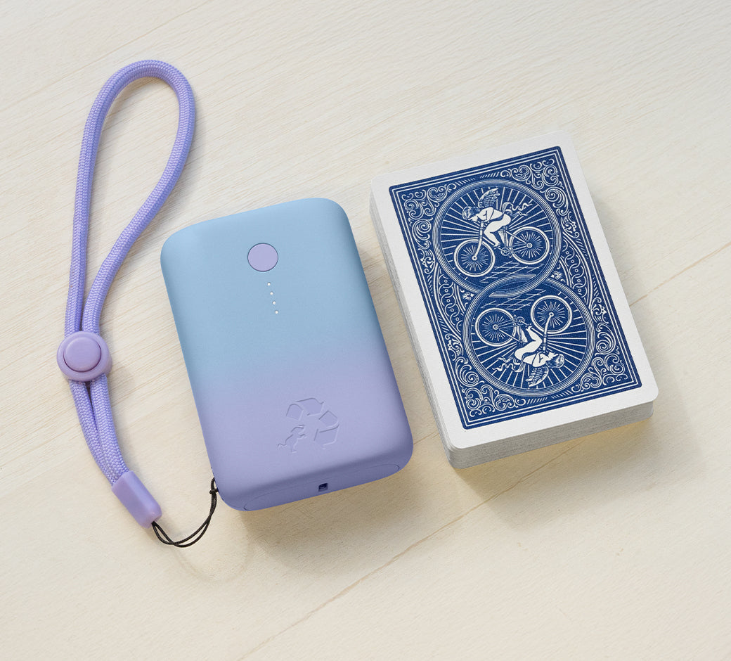 A lavender power bank with wrist strap beside a deck of blue Bicycle playing cards on a wooden surface.