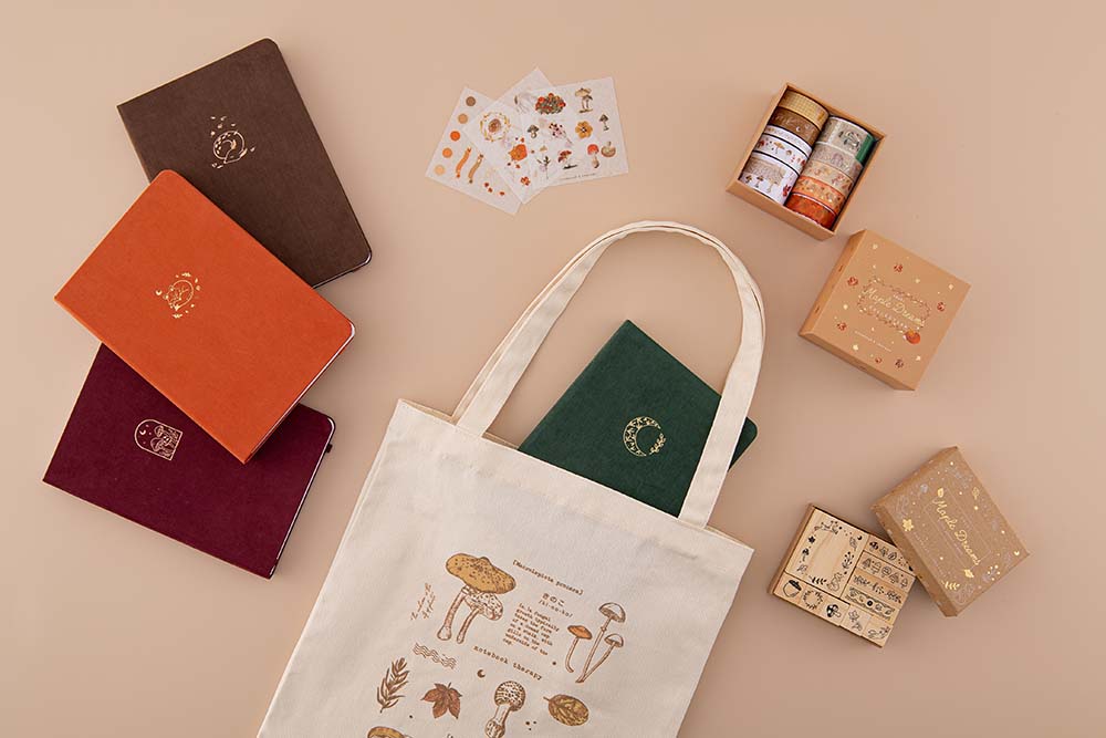 Tsuki ‘Kitsune’ Limited Edition Fox Bullet Journal with Tsuki ‘Kinoko’ Limited Edition Bullet Journal and Tsuki ‘Nara’ Limited Edition Bullet Journal and Tsuki ‘Maple Dreams’ Washi Tape Set and Tsuki ‘Maple Dreams’ Bullet Journal Stamp Set with Tsuki ‘Midnight Garden’ Limited Edition Bullet Journal inside Tsuki ‘Vintage Kinoko’ Tote Bag on beige background